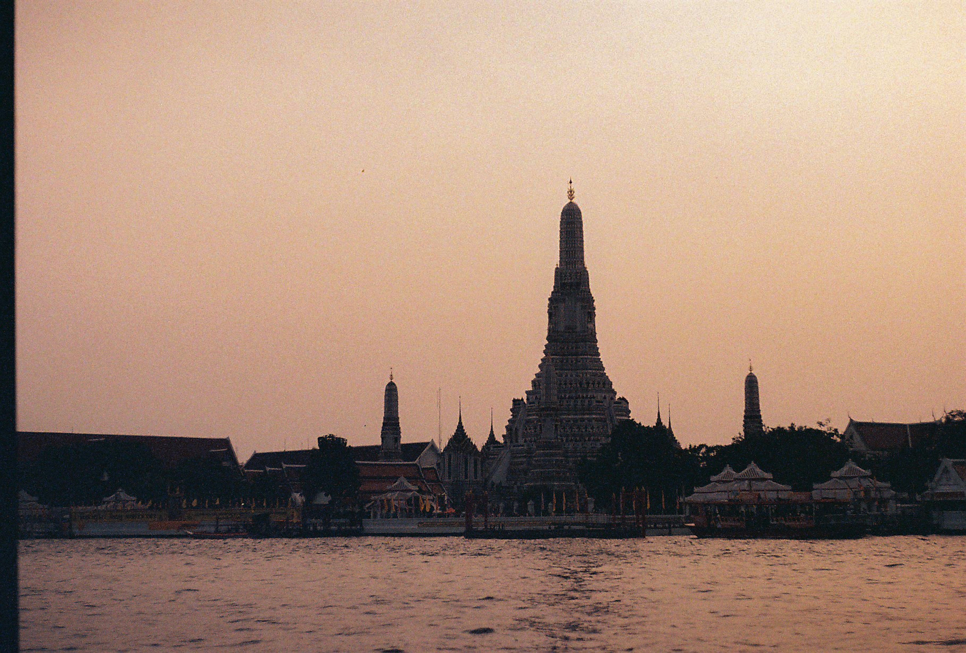 Wat Arun silhouetted across Bangkok’s Chao Phraya River beneath a pale peach evening sky