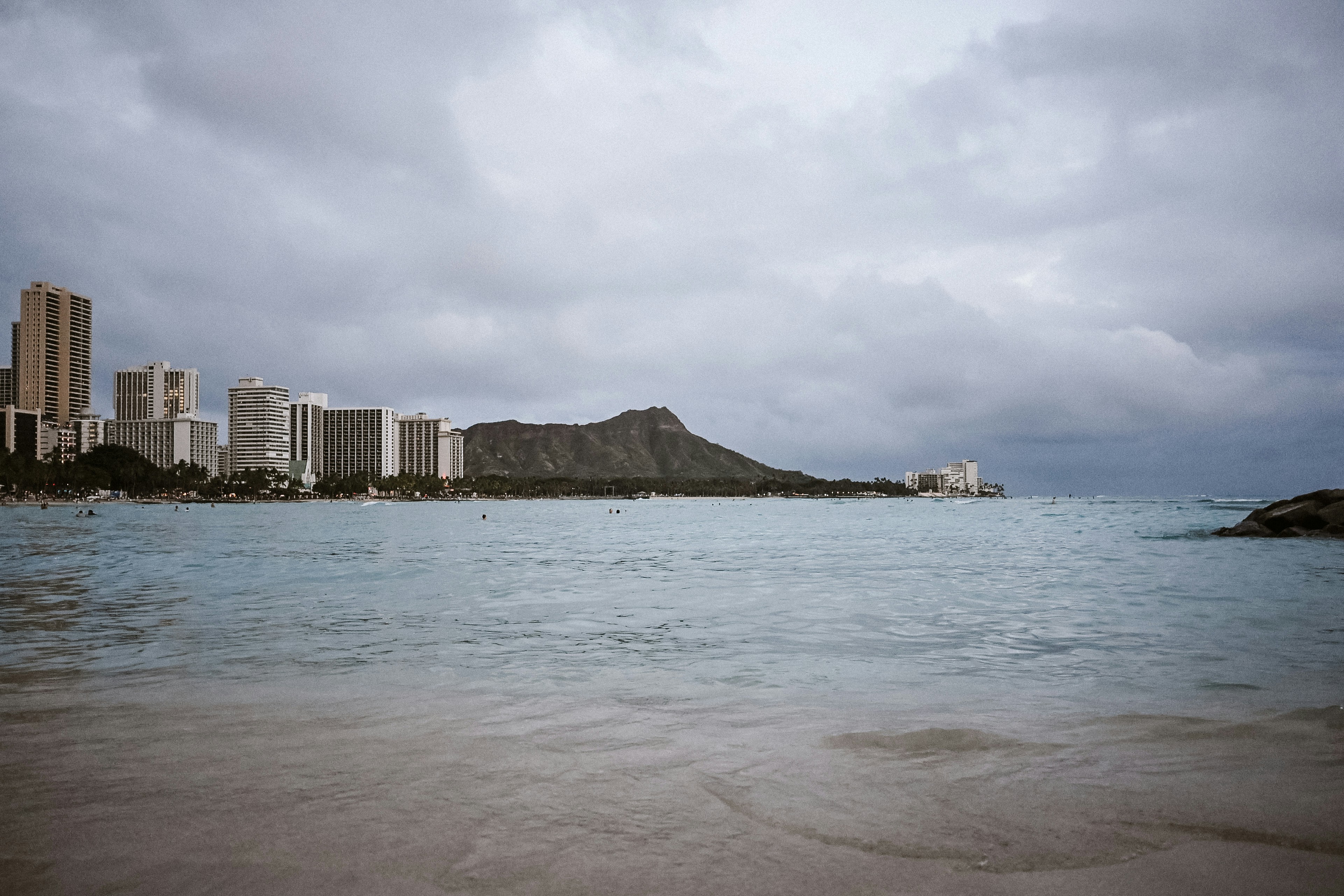 Waikīkī waterfront and the profile of Diamond Head beyond pale blue water under a cloudy sky