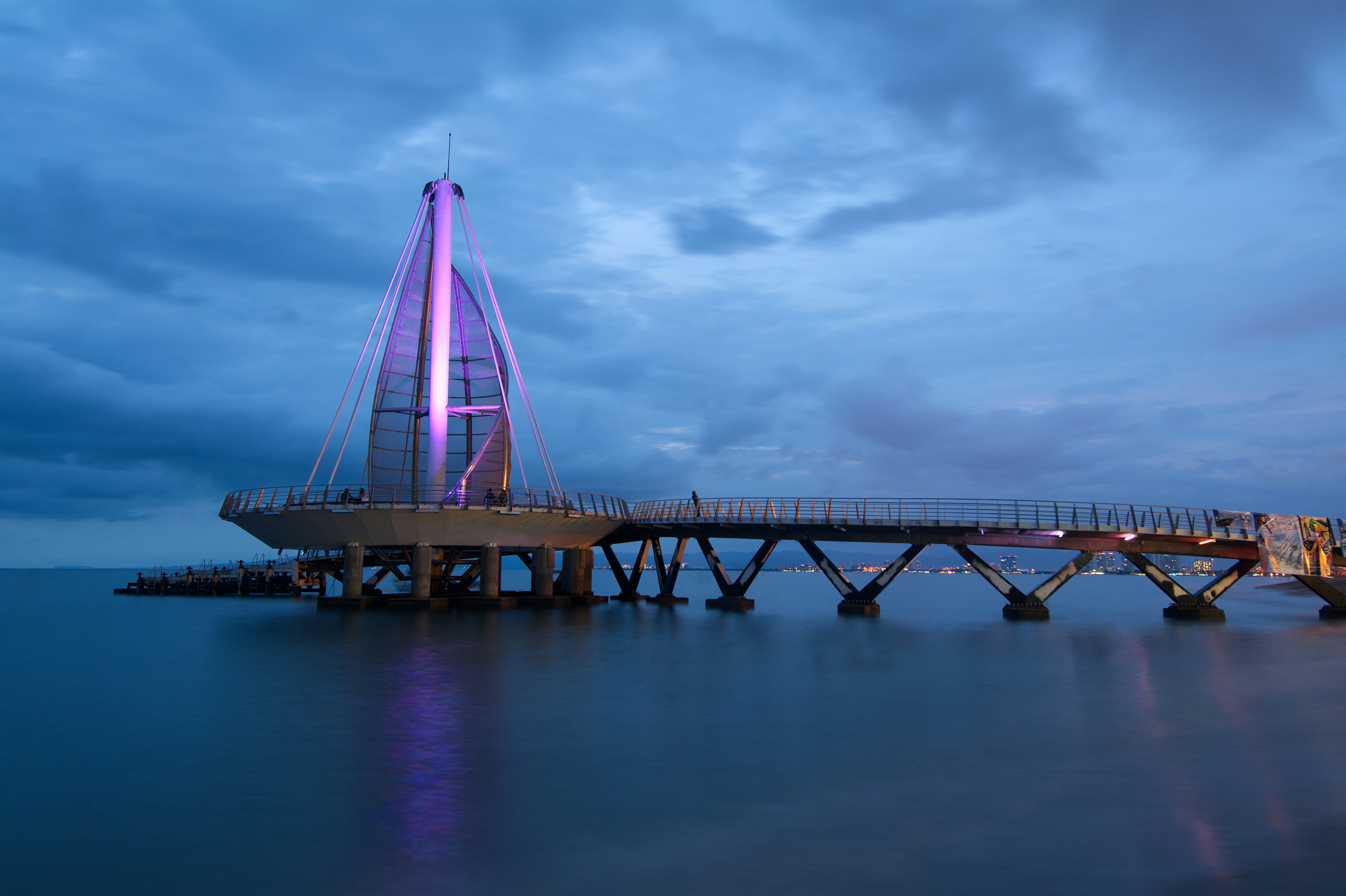 The sail-shaped Los Muertos pier illuminated in purple above Banderas Bay at dusk
