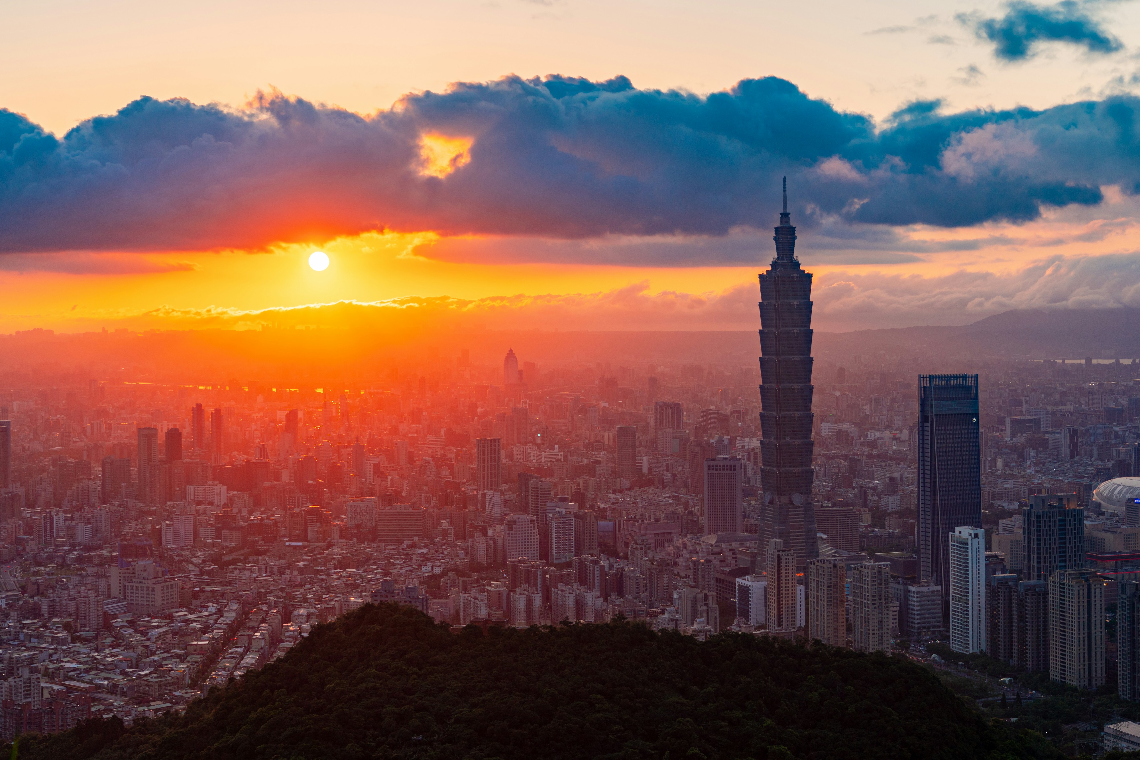 Taipei 101 rising above dense city blocks and wooded hills beneath a sunset sky